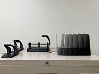 View of four black hole punches and a black desk organizer arranged on a white surface.