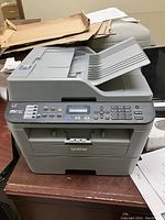 Front angle view of gray Brother multifunction laser printer with automatic document feeder on top and control panel with buttons and display on front.