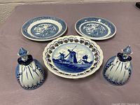 Set of two small blue and white floral plates, one larger scalloped plate with windmill design, and two ceramic figurines of women in traditional Dutch dress.