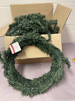 Artificial green wreath in a cardboard box, showing full circular shape and foliage texture.