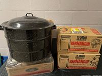 Black speckled enamel water bath canner with lid, two sealed boxes of Bernardin Mason jars stacked to the side.