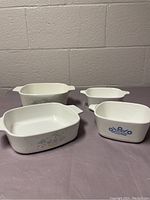 Lot of 4 Corning Ware casserole dishes on gray and pink fabric background: one blue cornflower design, one smaller multi-color flower pattern, two plain white dishes, all with two handles