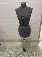 Full view of gray fabric covered dress form on chrome metal tripod stand against cinderblock wall.
