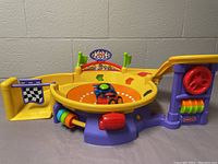 Fisher Price Lil Zoomers Spinnin Sounds Speedway race track with one blue and green race car on the circular track, showing side view with interactive parts and flags.