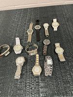 Top view of 12 assorted wristwatches laid out on a textured dark surface showing different band materials and watch faces.
