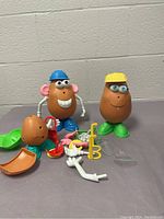 Lot of two assembled Mr. Potato Head figures along with many separate accessories placed in front of a gray cinder block wall.