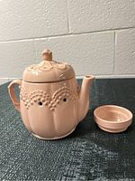 Front angled view of the pink ceramic teapot warmer with lid slightly raised and small dish next to it.