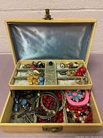 Jewelry box open showing the blue satin interior lid with star-like designs, green velvet ring slots, and assorted costume jewelry including earrings, rings, bracelets, and necklaces in various colors and materials.