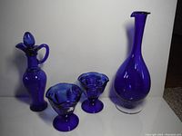 Full view of four cobalt blue glass items: one decanter with stopper and handle, two small ruffled edge bowls, and one tall narrow-neck vase with flared rim on clear base.