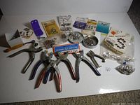 Full overview of the lot showing six pliers, small metal hardware components including grommets, snaps, and fasteners in various packaging and loose, displayed on white surface.