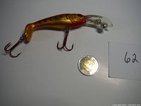 Side view of the gold-colored walleye crankbait lure with a clear diving lip and two red treble hooks next to a coin for scale.