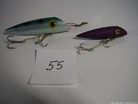 Photo showing two Luhr Jensen J-Plugs fishing lures: one larger blue-green with black spots, and one smaller purple lure, with a note '55' in front.