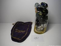 Tall translucent yellow vase filled with various large marbles. Purple Seagrams drawstring pouch placed next to vase. Numeral card '151' placed in front.