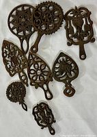 Nine antique cast iron trivets arranged on a white surface, various shapes and intricate patterns visible including floral and geometric designs.