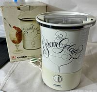 Overall view of the Girmi Gran Gelato electric ice cream maker with original box displaying product and branding. White cylindrical appliance with clear lid.