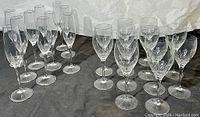 Grouped set of 15 clear signed crystal stemware glasses on dark cloth background. Includes both champagne flutes and wine glasses with intricate cut crystal patterns near base of bowl.
