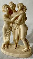 Front view of the small alabaster-resin sculpture depicting the Three Graces, showing the classical poses and delicate details of the three intertwined female figures on an oval base.