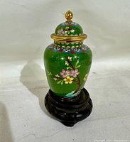 Front view of green cloisonne ginger jar with floral design, domed lid with gold finial, displayed on intricate dark carved wooden plinth base.