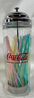 Front view of glass and chrome Coca-Cola straw dispenser holder filled with multicolor plastic straws. Clear cylindrical glass with Coca-Cola logo and 'Delicious and Refreshing' text.