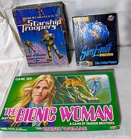 Three vintage game boxes displayed together: The Bionic Woman board game, Starship Troopers board game, and SimEarth PC game.