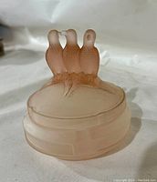 Side view of the pink frosted powder jar lid showing three detailed lovebirds perched on top, frosted glass surface visible.