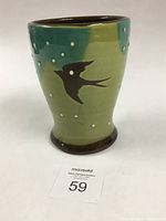 Front angled view of ceramic tumbler showing green and teal slip glaze, black bird stencil, white dot decoration, and glossy finish.