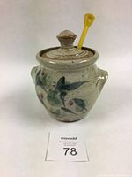 Photo of honey pot with lid, blue floral design, yellow honey wand in indent on lid, showing front and handles.