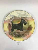 Full view of the circular ceramic plate featuring a black Scottie dog painted against a background of the Highlands with a cloudy sky and natural grasses.