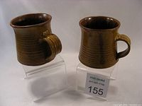 Two brown ceramic mugs with ribbed textures and round handles displayed on clear stands. Both mugs have a glossy glaze finish and no chips or cracks visible.