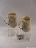 Two cream-colored ceramic mugs with mouse sculptures on handles, displayed on acrylic stands.