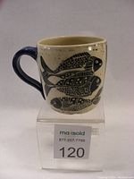 Front side of carved fish coffee mug showing three stylized fish in black on speckled cream background with blue handle
