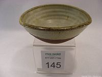 Ceramic bowl by Roger Stribley, displayed on a clear stand with Masold auction label visible. Shows top and side views highlighting glaze and unglazed base.