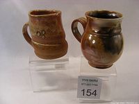 Two anagama style wood-fired coffee mugs displayed side by side on clear stands, showing different shapes and glaze patterns.