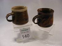 Two brown pottery mugs on clear stands showing front and side views to display shape and glaze