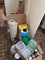 Box containing various household items including a rolled rug, baskets, canning jars, and boxes with supplies