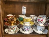 Set of 13 assorted teacups and saucers in various floral, gold, and pastel designs, stacked in two piles on a wooden surface.