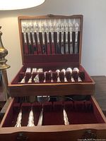 Wooden case open to show 12 decorative-handled dinner knives in upper section and various forks and spoons in lower sections separated by red velvet lining