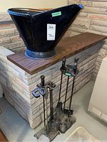 Photo showing a black metal ash bin with handle placed on bench, two fireplaces tool stands with tools leaning against stone fireplace