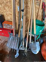 Cluster of wood-handled garden tools leaning against shed wall including rakes, forks, hoe, shovel, hammer