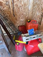 Four gas cans of different sizes, some with attached yellow spouts, assorted garden cages stacked vertically, red milk crate and garden hand tools visible in shed corner