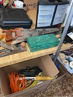 Four wooden hand saws with wooden handles, wooden hand planer, and a green metal box on a wood grain countertop, with a box of extension cords below.