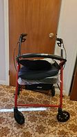 Red Drive rollator walker with black seat cushion and two memory foam cushions stacked on seat, positioned on carpeted floor against wooden door.
