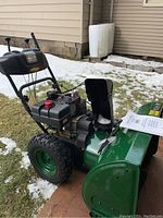 Side angle view showing green steel auger housing, black fuel tank with red cap, black control handles, operator’s manual on top