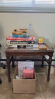 Front view of vintage wooden table with stacked board and card games on top, cardboard box with game pieces underneath.