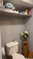 Shelf with bath towels, cleaning cloths, cleaning solutions, roll of paper towels, and spray bottles above a toilet with a wooden toilet paper stand on the floor next to it.