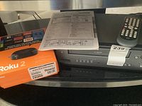 Magnavox VCR device with remote on top, next to the orange Roku 2 box with cables and instruction manual visible.