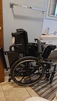 Full side view of the black Invacare Patriot wheelchair placed on tiled floor showing its large rear wheels and padded armrests.