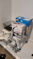 Shot showing two white shower stools with blue legs, black walker basket, OttLite lamp box, and AquaSense raised toilet seat box on top of a shower stool. Chrome metal handrails seen in front.