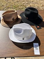 Three brand new hats arranged on a table outdoors: one white perforated fedora, one brown perforated woven fedora with brown band, and one black felt fedora with gray band and button.