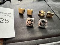 Three pairs of cufflinks displayed together on a black cloth background. Includes gold-tone textured square pair, gold-tone rectangular pair with inset, and silver-tone square pair with black circle center.